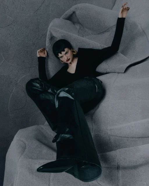 Fashion shot of a woman with short dark hair in black top, pants, and boots sitting on a gray fabric surface, looking cool and collected.