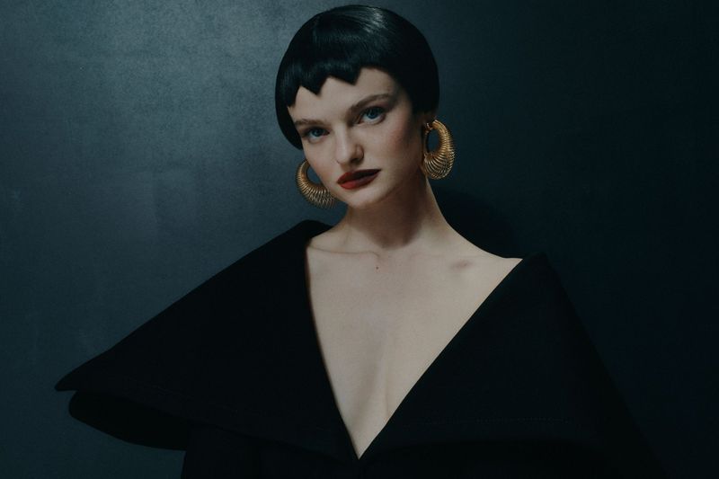 Glamorous beauty portrait: adult woman with black bowl cut, red lipstick, wearing gold spiral earrings and a black dress, studio shot.
