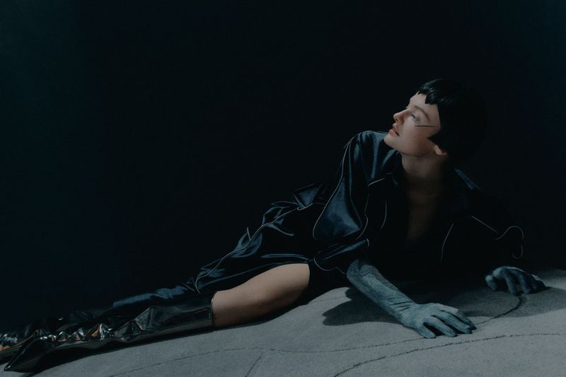Woman in a satin robe with leather boots poses in a dimly lit, moody fashion shot with cool tones on a textured surface.