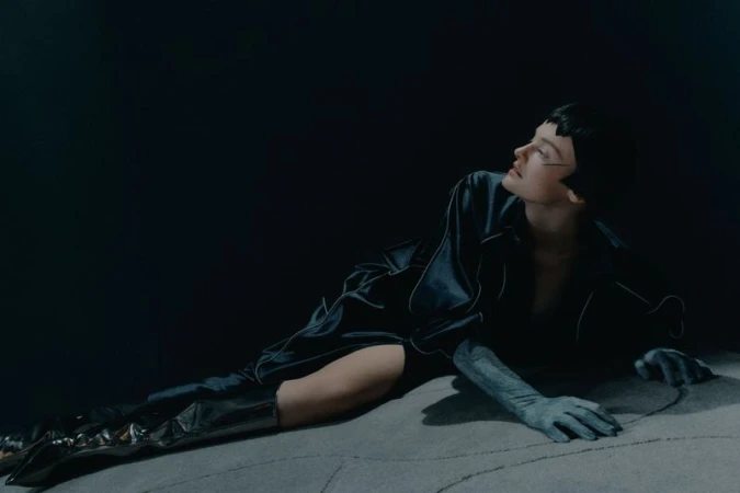 Woman in a satin robe with leather boots poses in a dimly lit, moody fashion shot with cool tones on a textured surface.