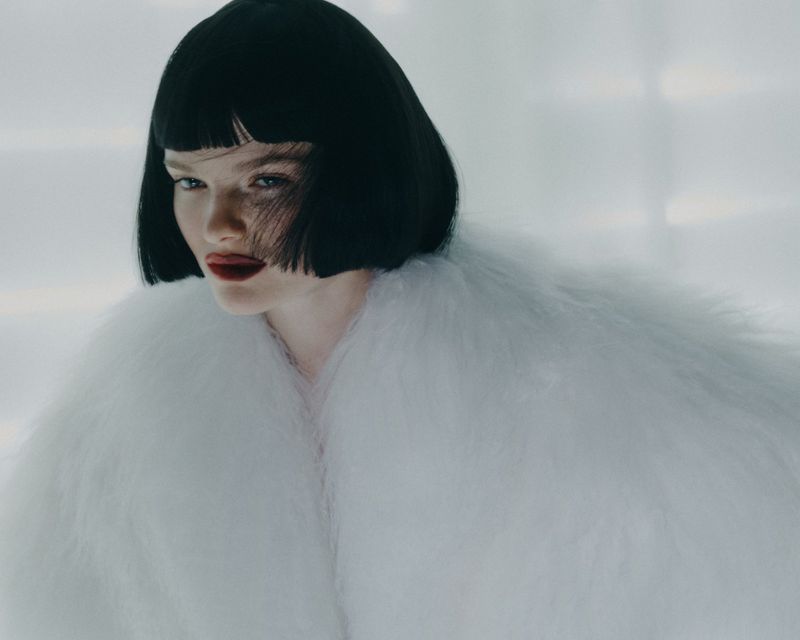 Portrait of a woman with a bob haircut wearing a white faux fur coat against a soft white background in a beauty fashion shot.