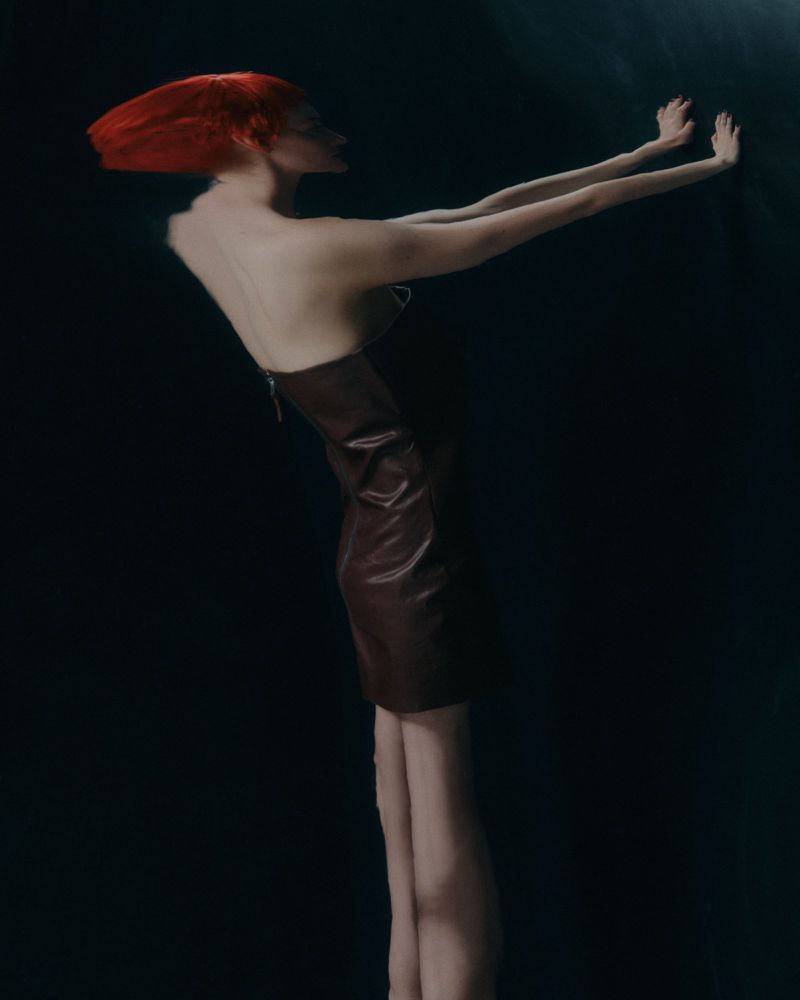 Artistic studio portrait of a person with red hair wearing a dress, posing with arms extended in front of a dark background. 
