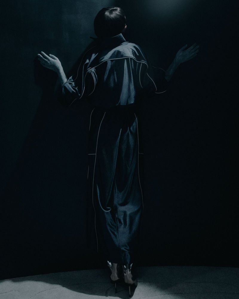 Back view of a person in dark blue outfit with white trim, arms outstretched, against a shadowy blue wall. Lower portion visible.