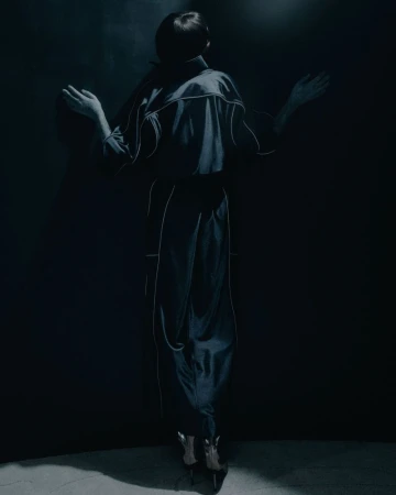 Back view of a person in dark blue outfit with white trim, arms outstretched, against a shadowy blue wall. Lower portion visible.