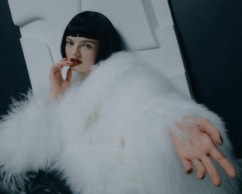 Portrait of a woman with black hair wearing a white, fluffy coat indoors, looking at the camera with hand near her face.