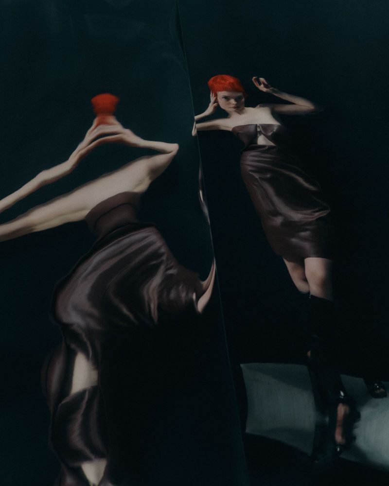 Elegant woman in a dark dress and red hair poses in a surreal studio environment, reflected in a distorted mirror with dark color grading.