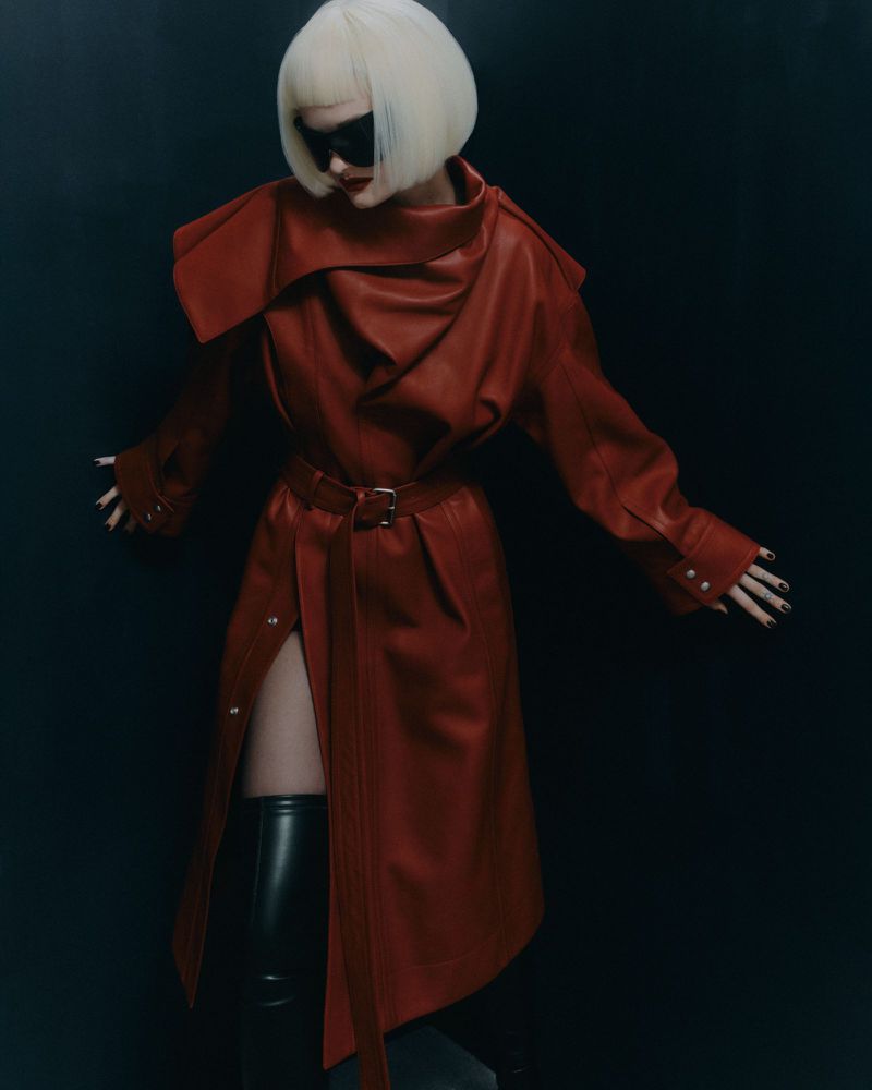 A stylish adult female posing in a red belted coat with long sleeves and black thigh-high boots against a dark backdrop in a studio.