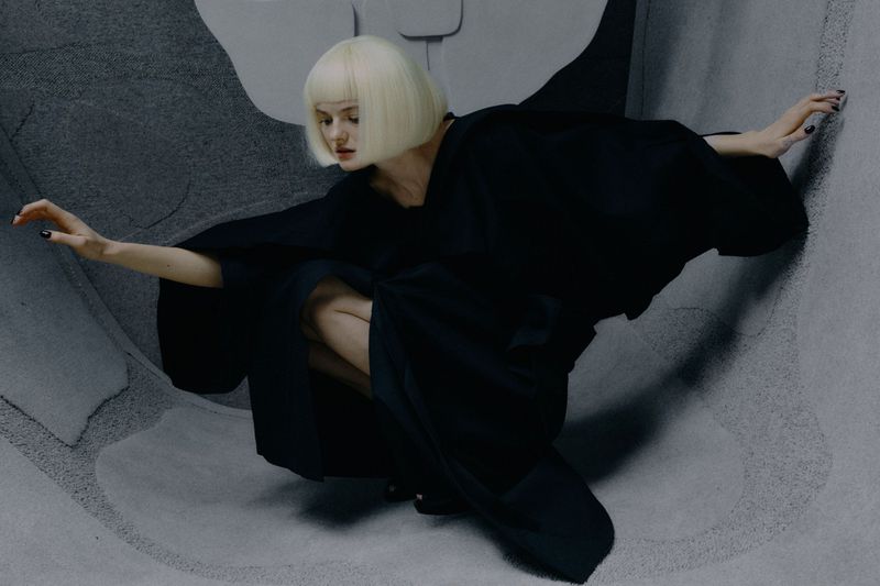 Woman in a black kimono crouching in a photo studio. She has platinum-blonde hair and fair skin. The photo studio backdrop is grey.