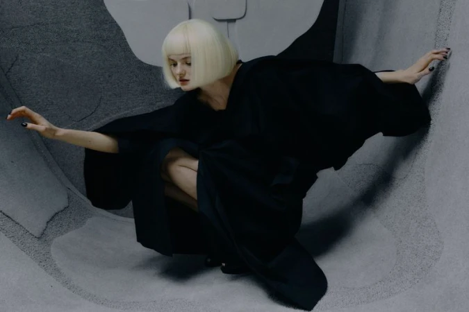 Woman in a black kimono crouching in a photo studio. She has platinum-blonde hair and fair skin. The photo studio backdrop is grey.