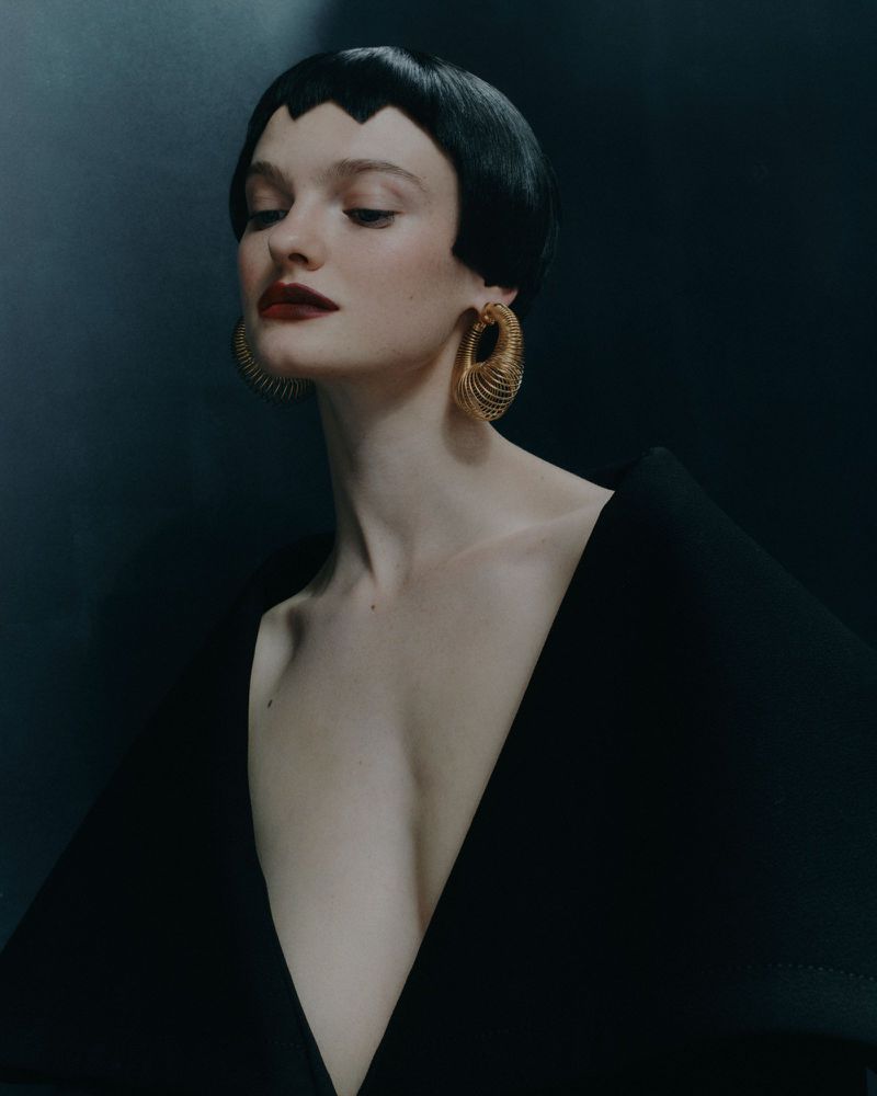 Portrait of a fair-skinned young woman with a black bob hairstyle wearing a black V-neck garment and gold earrings against a dark background.