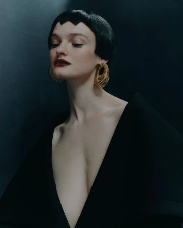 Portrait of a fair-skinned young woman with a black bob hairstyle wearing a black V-neck garment and gold earrings against a dark background.