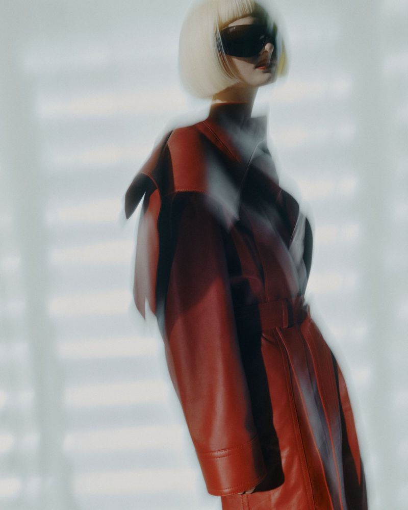 Model with blonde bob and dark sunglasses wears a red leather coat in a blurred studio setting with white horizontal lines in the background.