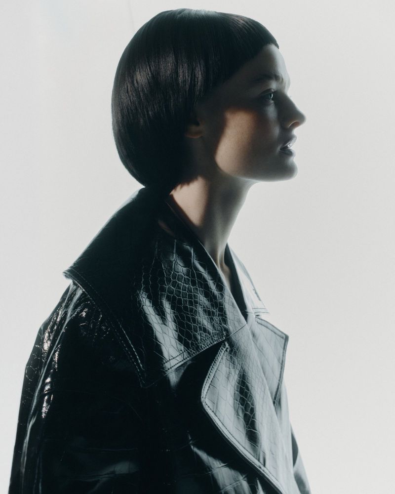 Portrait of a woman with dark bob hairstyle in a black faux snakeskin trench coat against a bright background in a fashion studio.