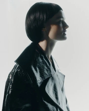 Portrait of a woman with dark bob hairstyle in a black faux snakeskin trench coat against a bright background in a fashion studio.