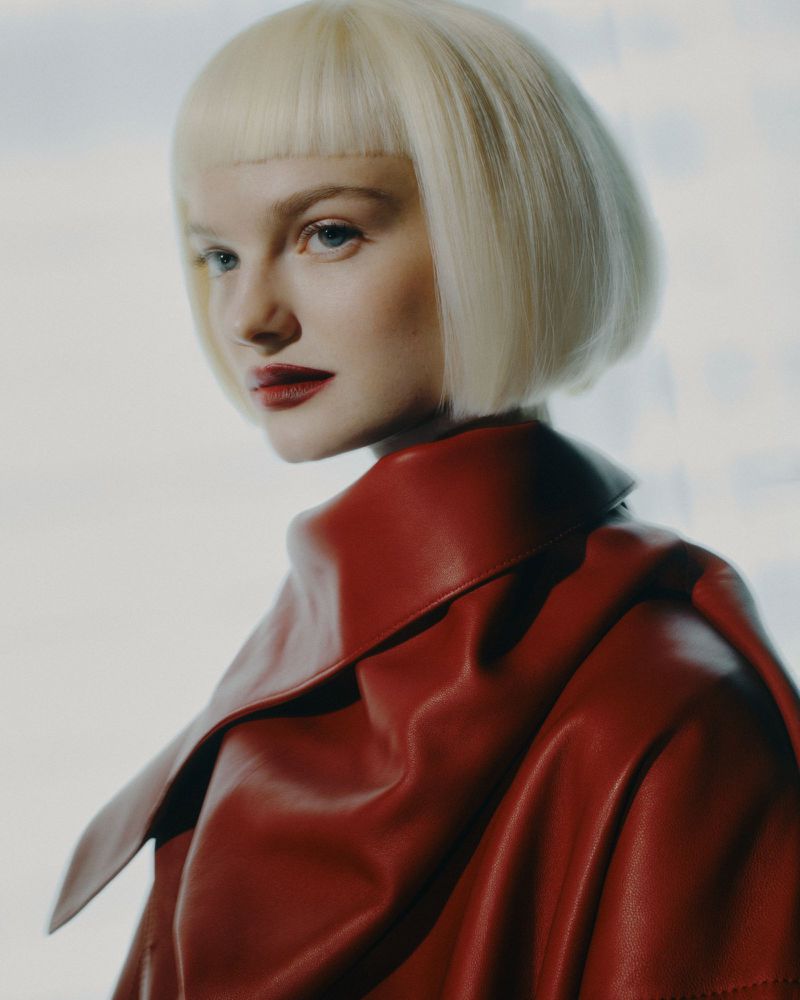 Close-up shot of a fair-skinned young woman with a short blonde bob haircut, red lipstick, and wearing a red leather coat.