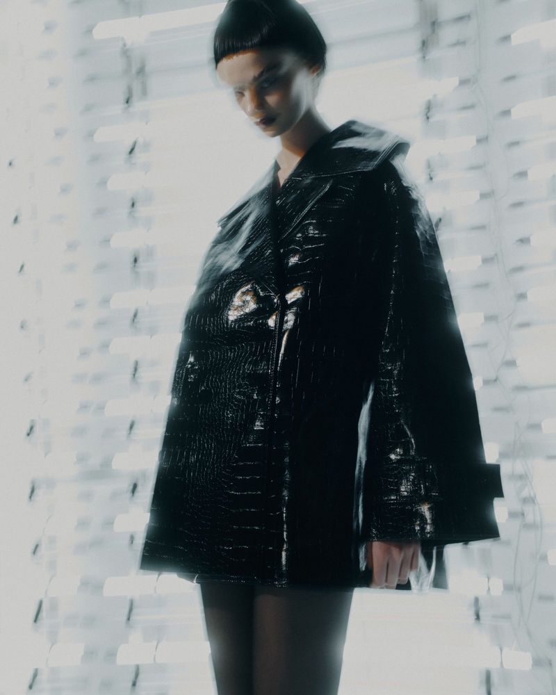 Fashion shot of a woman wearing a black shiny coat and a beanie standing in an indoor setting with blurry lights in the background.