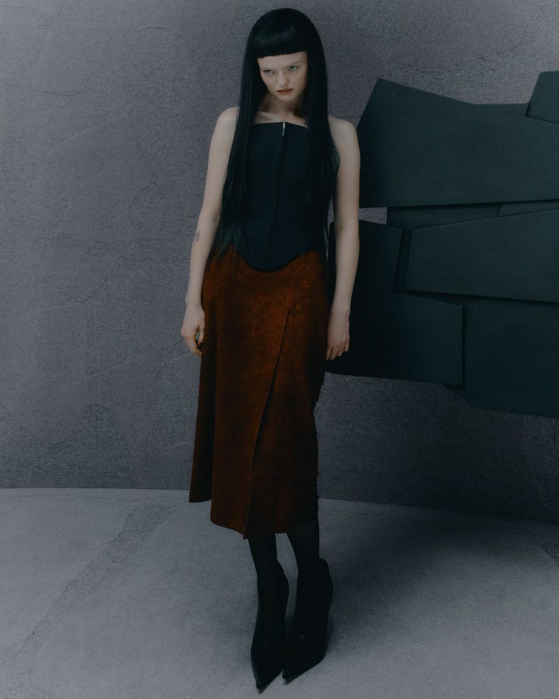 Young woman with bangs poses in a black top and rust-colored skirt against a gray wall with stacked dark geometric shapes.