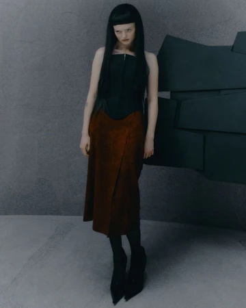 Young woman with bangs poses in a black top and rust-colored skirt against a gray wall with stacked dark geometric shapes.