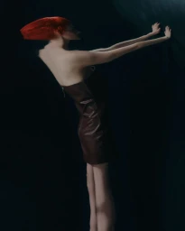 Model with vibrant red hair wearing a dark brown strapless dress pressing hands against a dark wall for Sicky Magazine.