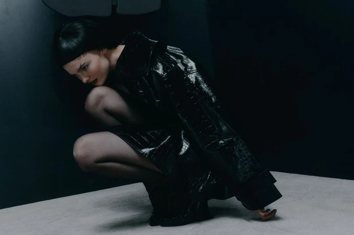 Model in a black glossy trench coat crouching against a dark studio background for Sicky Magazine