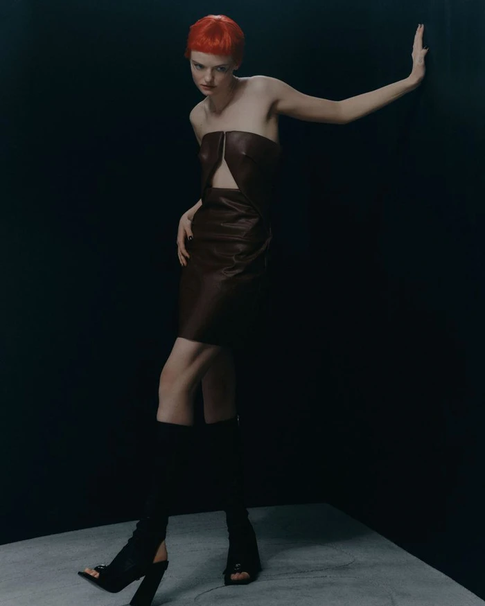 Model with bright red hair wearing a dark brown strapless leather dress leaning against a dark wall in a studio shoot for Sicky Magazine