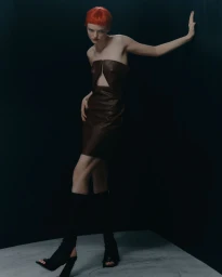 Model with bright red hair wearing a dark brown strapless leather dress leaning against a dark wall in a studio shoot for Sicky Magazine