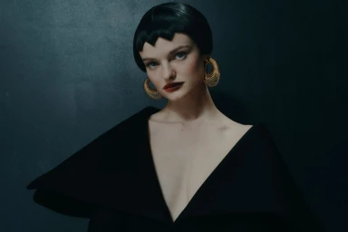 Model with dark geometric bob and gold hoop earrings wearing structured black top against a dark textured wall for Sicky Magazine.