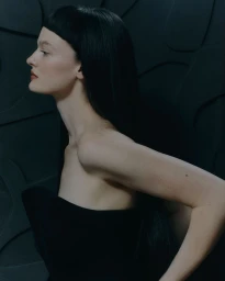 Profile portrait of a woman with dark hair and bangs wearing a black strapless garment against a dark patterned wall.