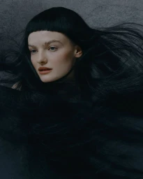 Close-up portrait of a woman with long black hair, heavy bangs, and dark flowing fabric against a textured grey background.