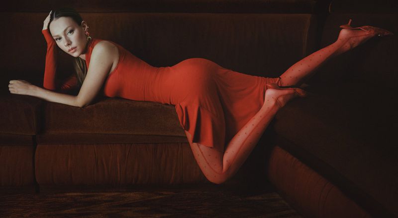 Ester Exposito lying on a dark velvet sofa wearing a red dress and patterned tights for Marine Serre