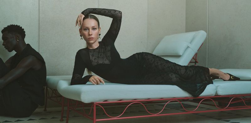Adult female model in long black sheer gown with crescent moon pattern, lying on a cushioned chaise lounge, studio fashion shot.