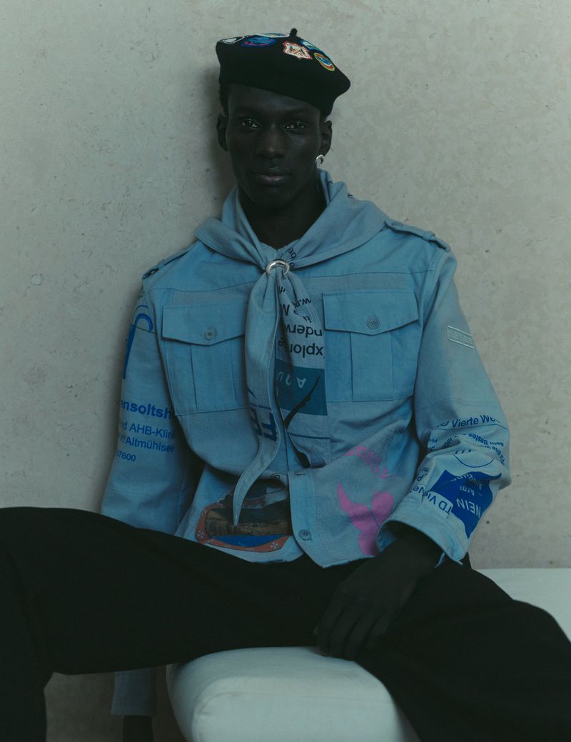 Model wearing a light blue graphic printed shirt and black beret seated against a neutral wall