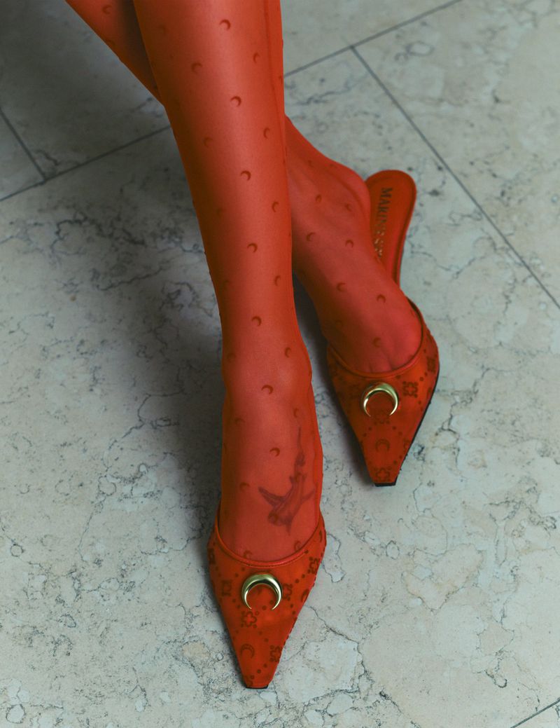 Close-up of legs wearing orange patterned tights and red pointed-toe mules with gold hardware against a light stone floor