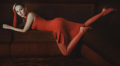 Ester Exposito lying on a dark velvet sofa wearing a red dress and patterned tights for Marine Serre