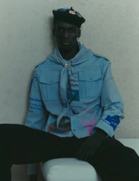 Model wearing a light blue graphic printed shirt and black beret seated against a neutral wall