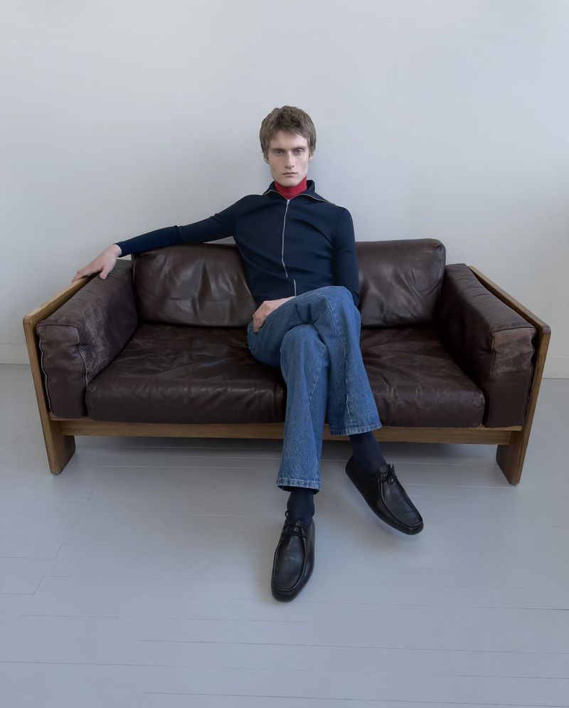 Model seated on a brown leather sofa wearing a dark zipped top and blue jeans against a light background.