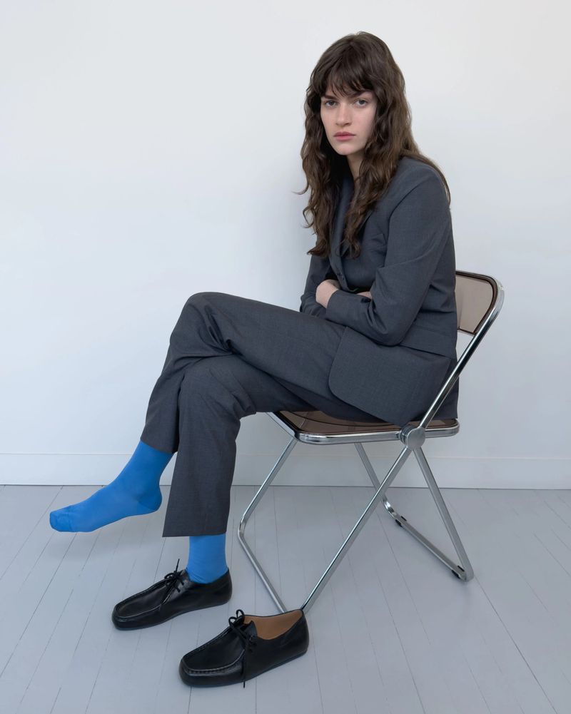 An adult female sits on a chair in a gray suit with one foot socked and one foot showing shoes, against a plain white wall.