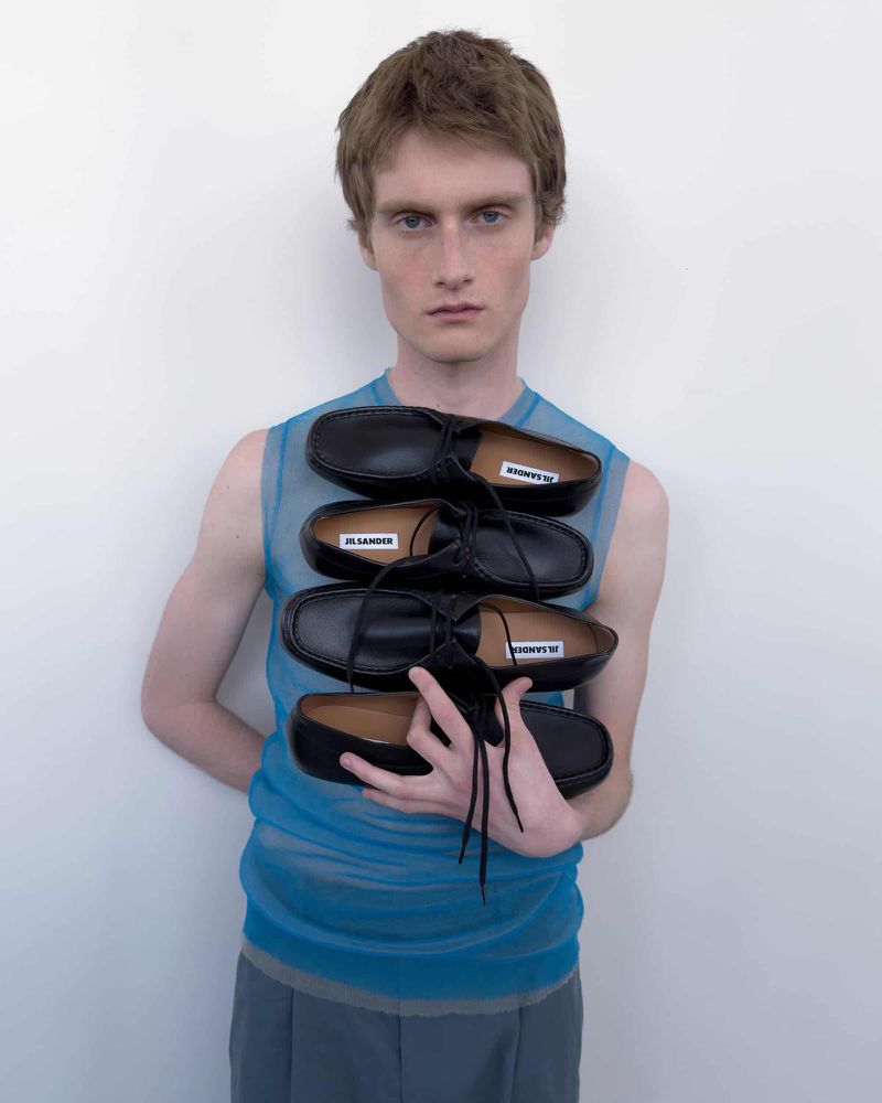 Model wearing blue sheer top holding a stacked column of black leather shoes against white wall