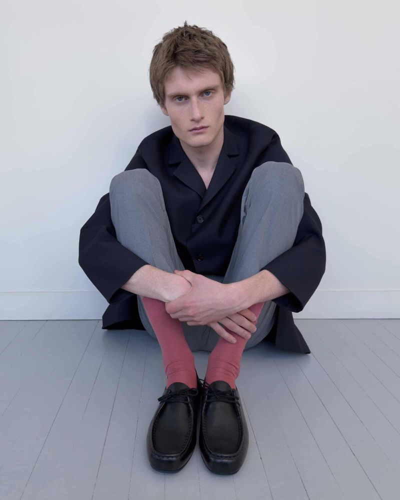 Young male model sitting on a grey floor against a white wall wearing a dark jacket, grey trousers, pink socks, and black shoes.