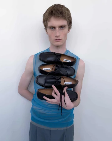 Model wearing blue sheer top holding a stacked column of black leather shoes against white wall
