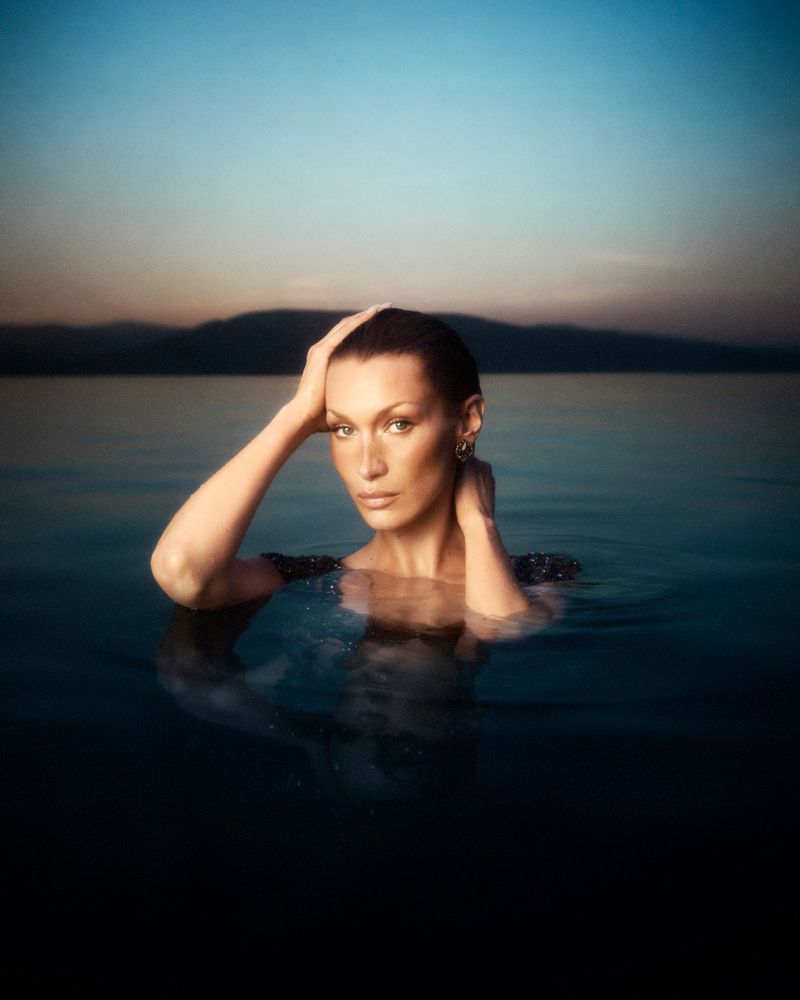 Bella Hadid partially submerged in calm water at dusk with a distant shoreline visible under a gradient sky