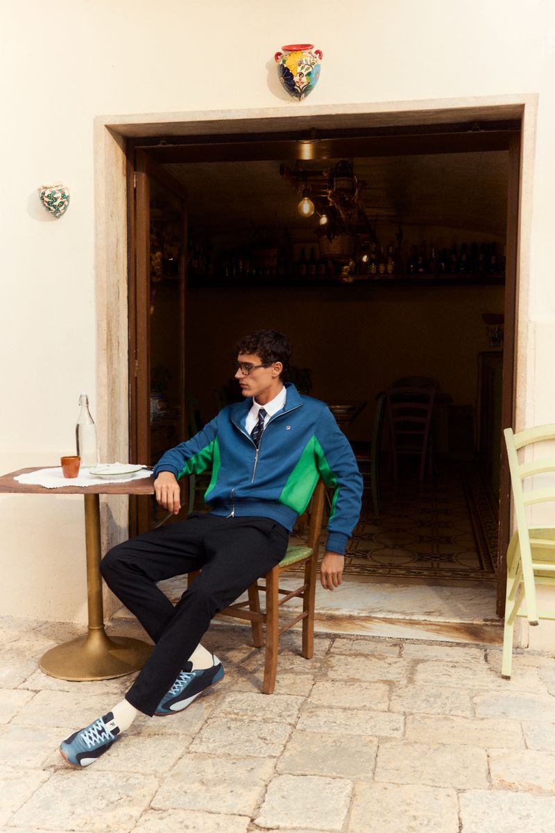 A man in a blue and green zip jacket seated at an outdoor cafe table for Fila