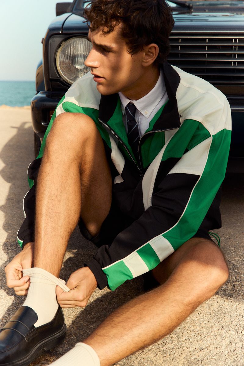 Model wearing a green, black, and white track jacket sitting by a car near the ocean