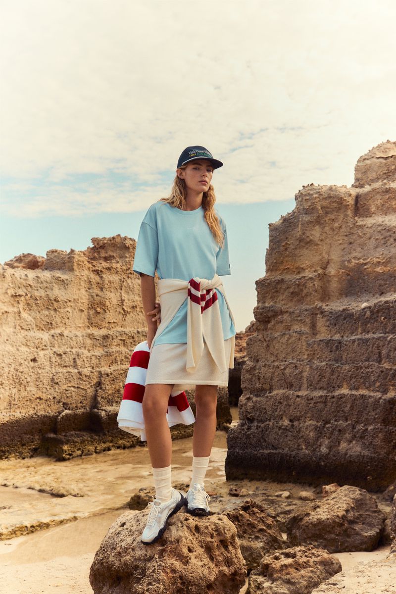 Model standing on rocky terrain wearing a light blue t-shirt, white skirt, baseball cap, and white sneakers for Fila Spring / Summer 26.