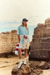 Model standing on rocky terrain wearing a light blue t-shirt, white skirt, baseball cap, and white sneakers for Fila Spring / Summer 26.