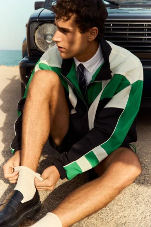 Model wearing a green, black, and white track jacket sitting by a car near the ocean