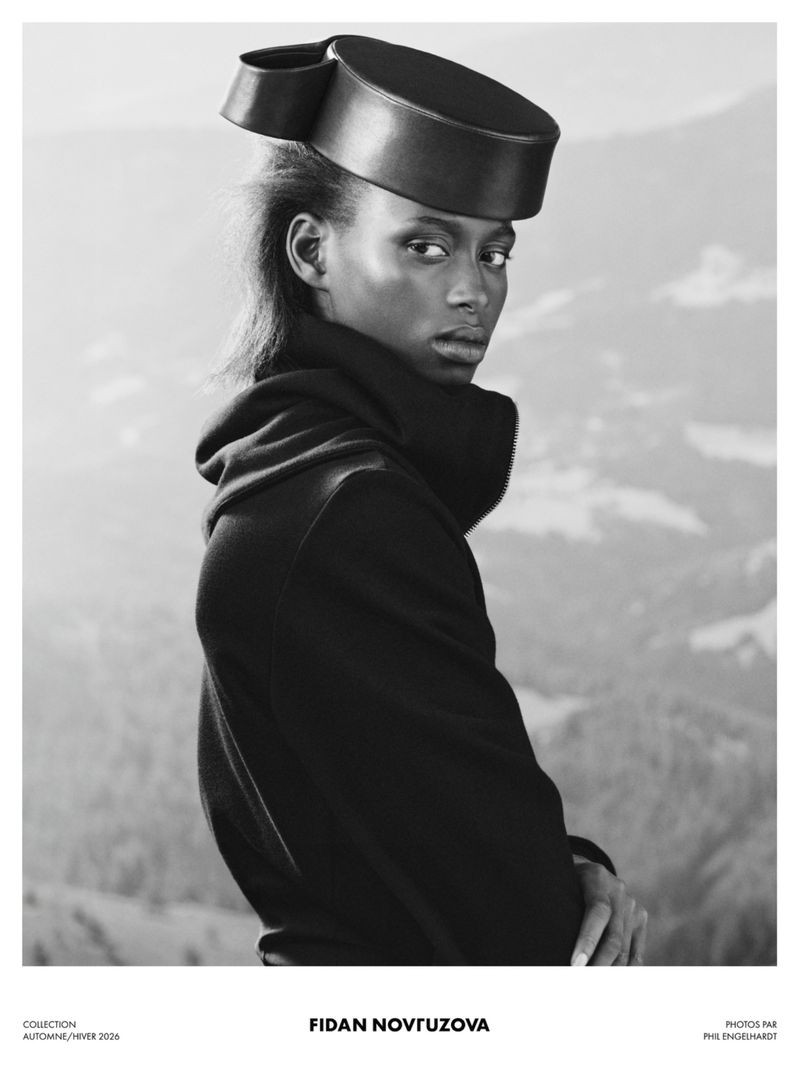 Model wearing a structured leather pillbox hat and dark turtleneck against an outdoor landscape background