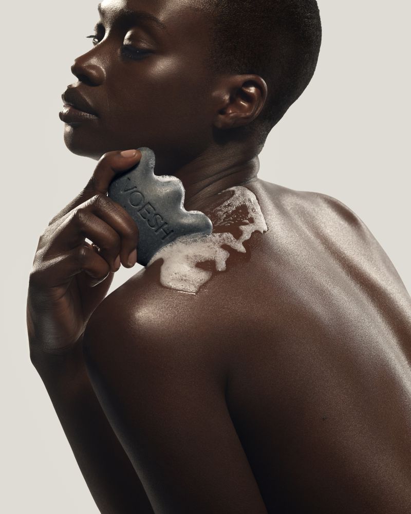 Black woman holding a charcoal cleansing bar with white lather on her shoulder against a neutral background for VOESH.