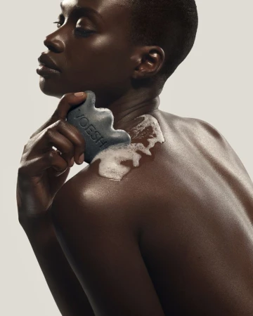 Black woman holding a charcoal cleansing bar with white lather on her shoulder against a neutral background for VOESH.