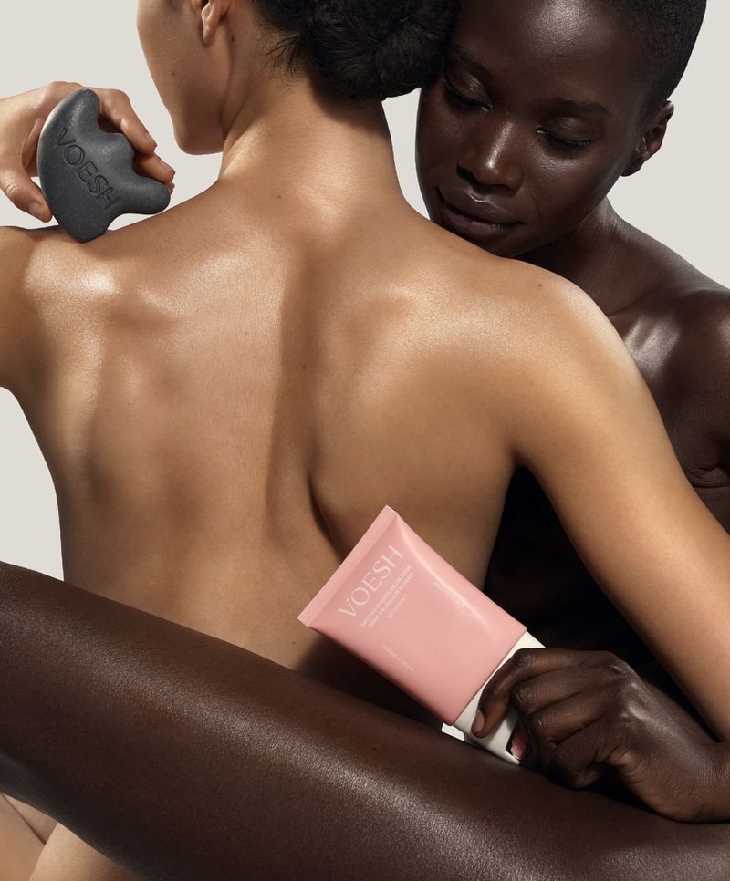 Two models embracing against a seamless background while holding a dark grey gua sha tool and pink skincare tube.